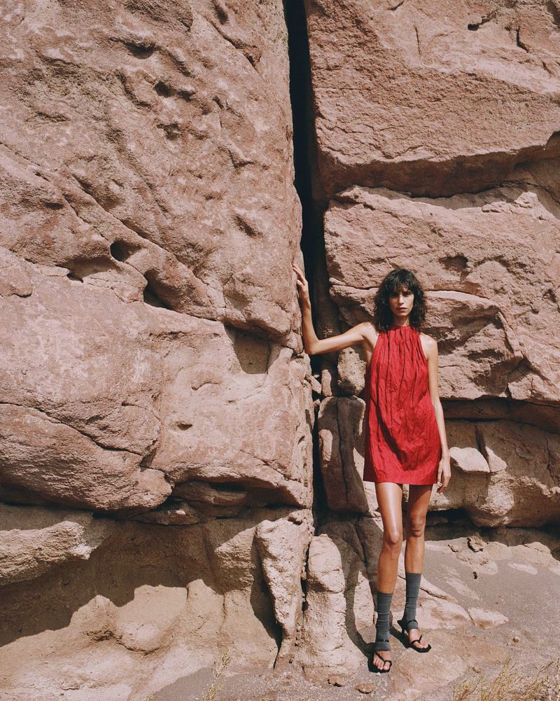 A woman in a red dress stands against a large rock formation in an outdoor setting, creating a striking contrast of color and texture.
