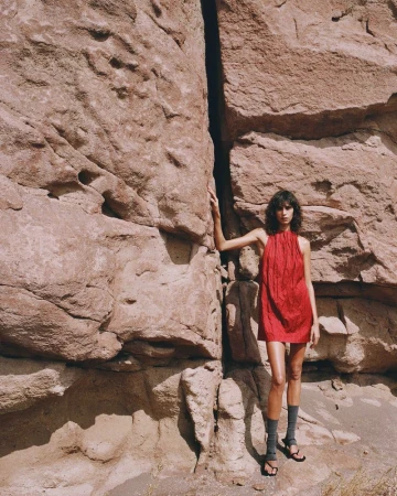 A woman in a red dress stands against a large rock formation in an outdoor setting, creating a striking contrast of color and texture.