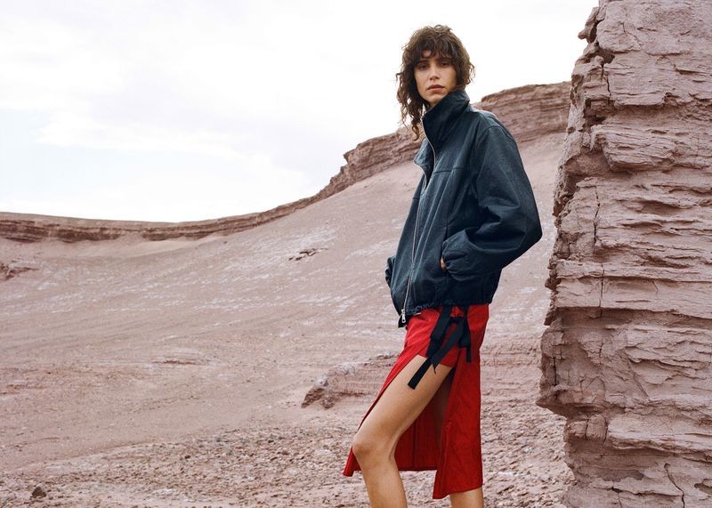 Woman in jacket and red skirt poses in a desert landscape with rock formations under an overcast sky for a fashion editorial.