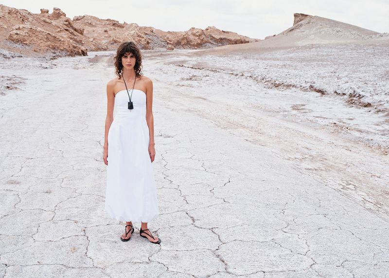 A woman in a white dress stands in a barren landscape with distant rock formations, creating a stark yet elegant fashion portrait.