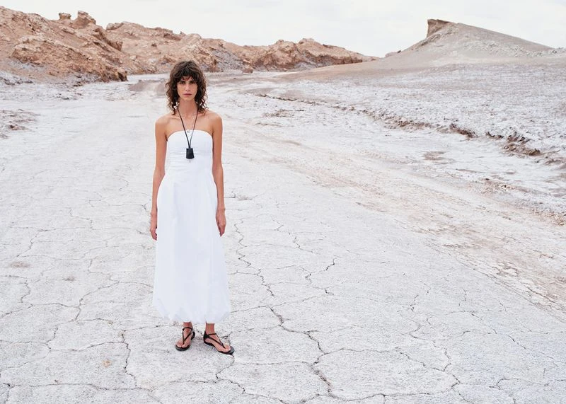 A woman in a white dress stands in a barren landscape with distant rock formations, creating a stark yet elegant fashion portrait.