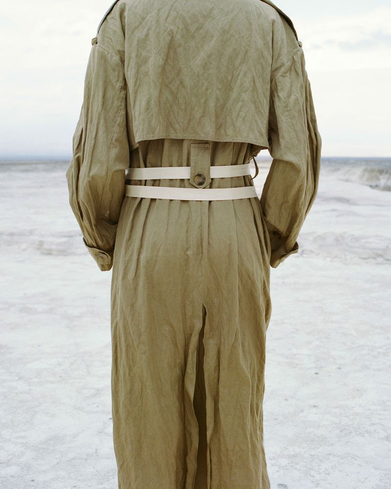 Back view of a person in a khaki trench coat with a white belt, standing on a sandy beach near the ocean waves under a cloudy sky.