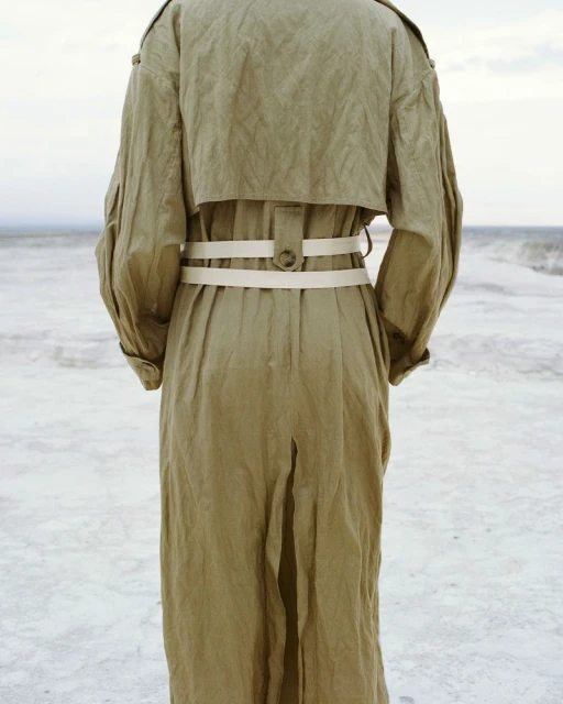 Back view of a person in a khaki trench coat with a white belt, standing on a sandy beach near the ocean waves under a cloudy sky.