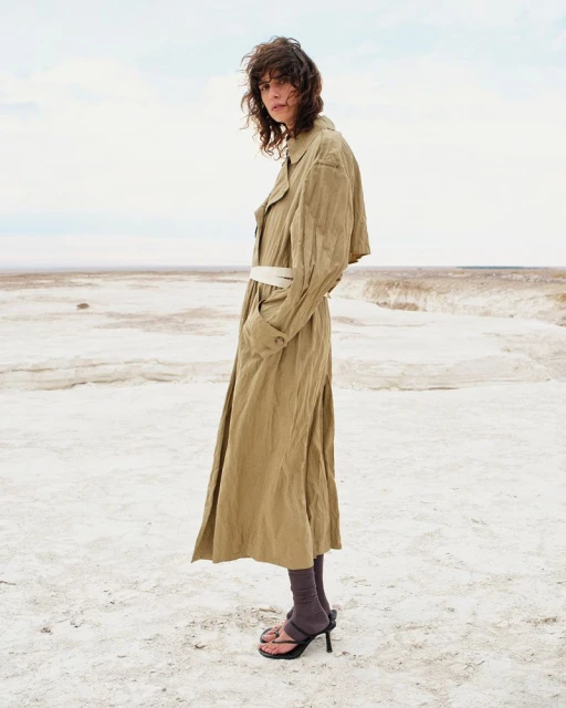 A woman in a tan trench coat standing on a cracked white ground beneath a bright overcast sky in a fashion editorial.