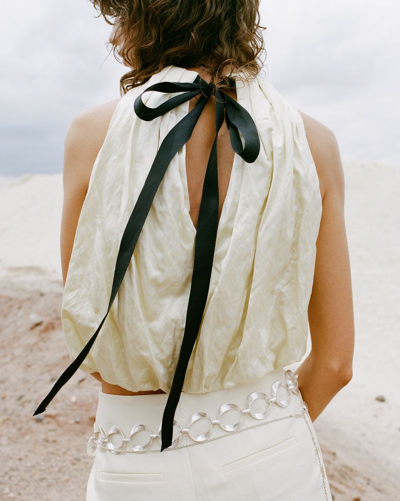 A woman with wavy brown hair wears a sleeveless cream top with a black ribbon tied at the neck, in an outdoor desert setting.