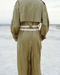 Back view of a model wearing an oversized khaki green trench coat with double cream waist belts against a pale sandy beach.