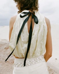 Model seen from behind wearing an ivory halter top with a black ribbon bow and white trousers against sand dunes.