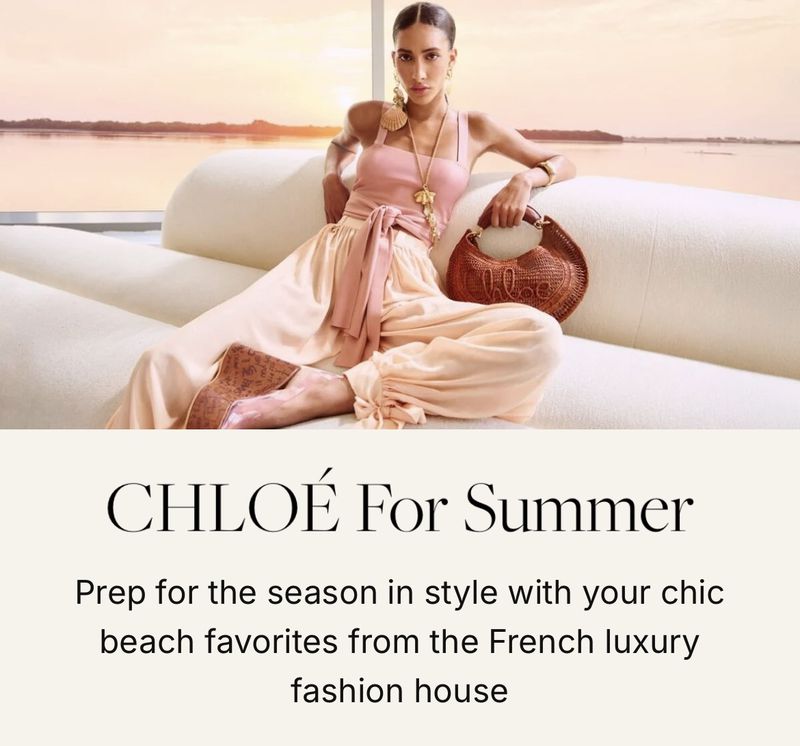 A woman sits on a white sofa near water in a fashion shot. She wears a pink top and light-colored pants, holding a round wicker bag.