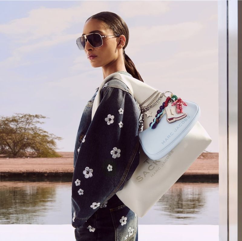 Model wears floral-embroidered denim jacket and carries Marc Jacobs tote bag in an outdoor setting near a body of water.