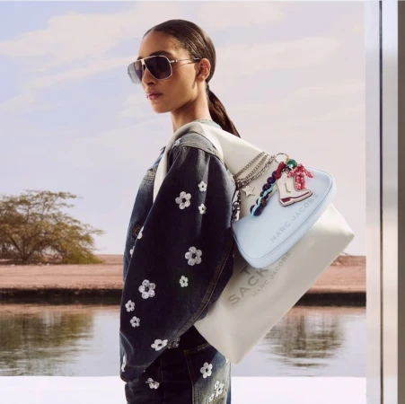 Model Titia Le Roux wearing aviator sunglasses, a flower-embellished denim jacket, and Marc Jacobs bags by a body of water.