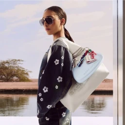 Model Titia Le Roux wearing aviator sunglasses, a flower-embellished denim jacket, and Marc Jacobs bags by a body of water.