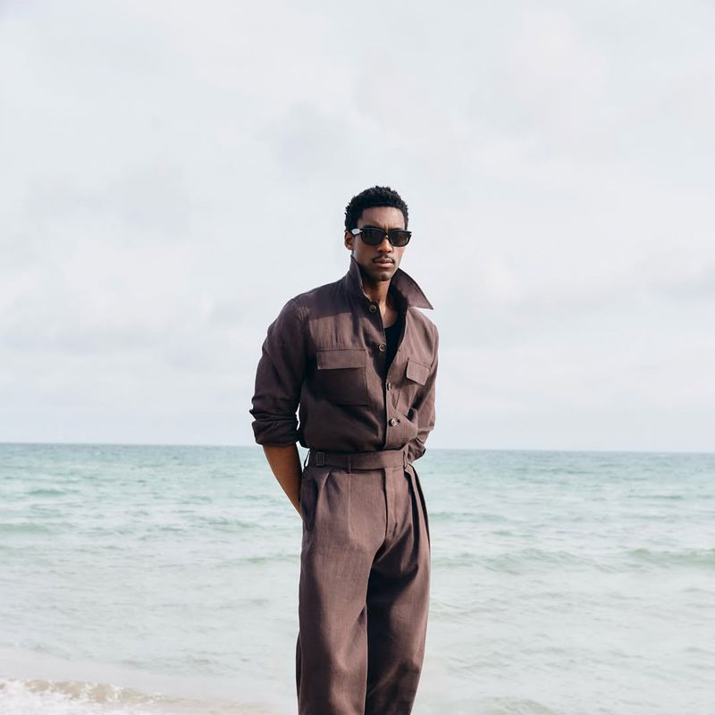 A man wearing sunglasses and a brown jumpsuit stands on a sandy beach with ocean and sky in the background in a medium outdoor shot.