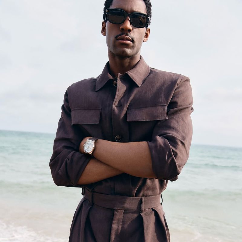 Dark-skinned adult male with a mustache poses with arms crossed on a beach while wearing sunglasses and a brown linen collared jumpsuit.
