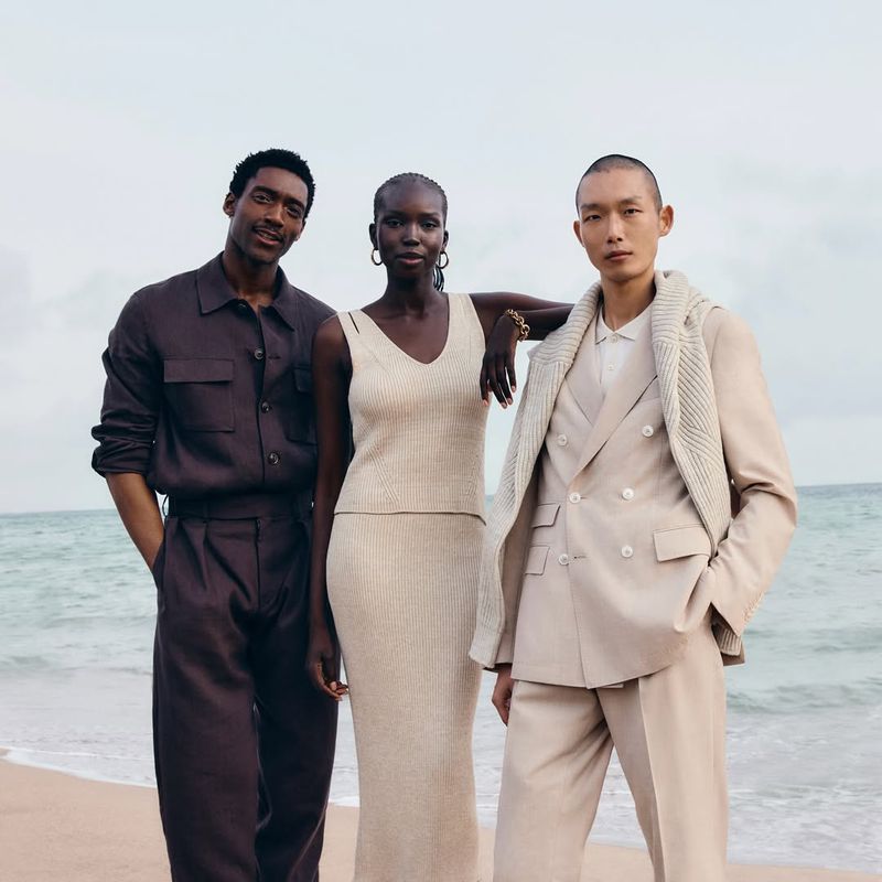 Three fashion models—two men and a woman—pose on a beach. The woman is centered between the two men, all wearing neutral-toned clothing.