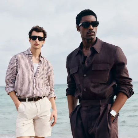 Two stylish men pose on a beach wearing sunglasses, button-down shirts, and shorts against a backdrop of the ocean and sky.