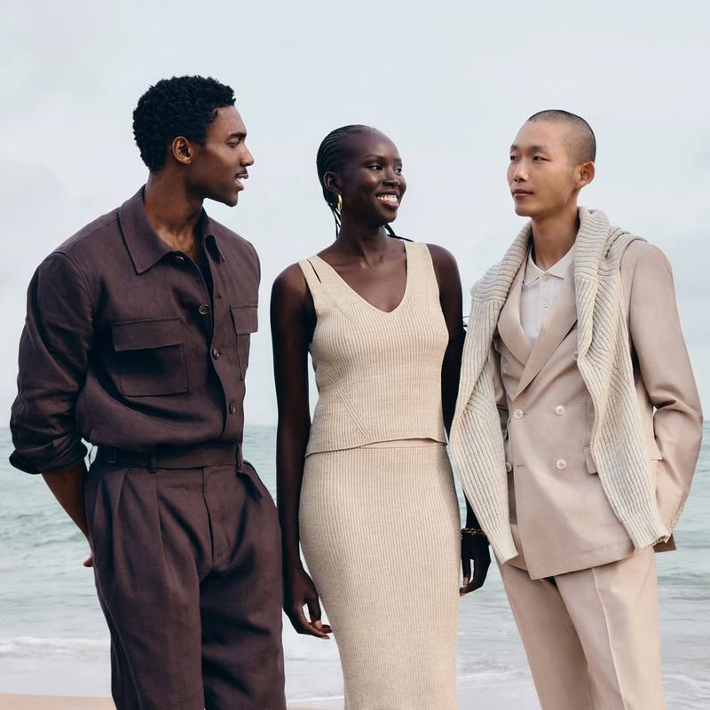 Three diverse fashion models pose on a sandy beach with the ocean in the background. One man wears brown, a woman wears cream, another man wears beige.
