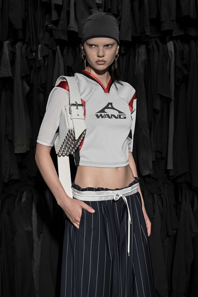 A fashionably dressed woman in a sports-inspired crop top, pleated skirt, and a beanie stands in front of a rack of black shirts.