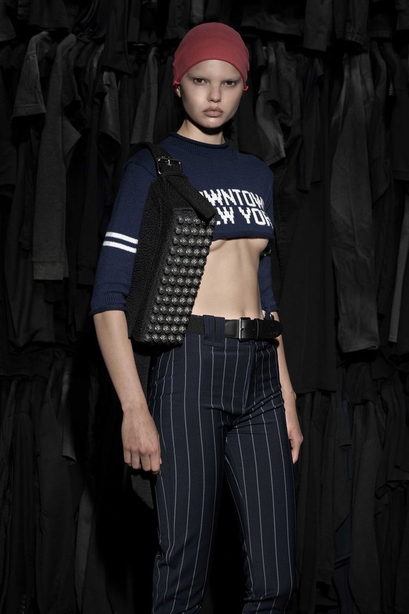 Fashion shot of a fair-skinned young adult woman posing indoors wearing a cropped top, pants, head wrap, and studded bag.