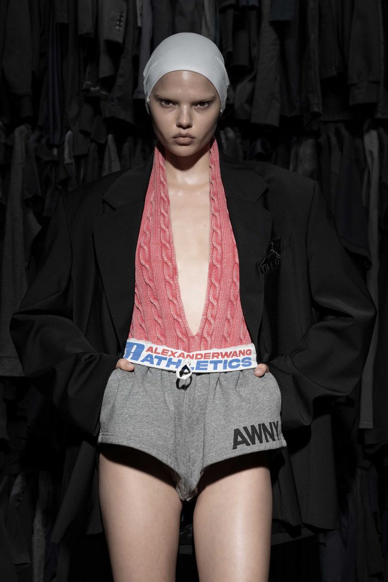 A woman in athletic shorts, sweater, and blazer poses against a backdrop of hanging dark clothes for a fashion-forward photograph.