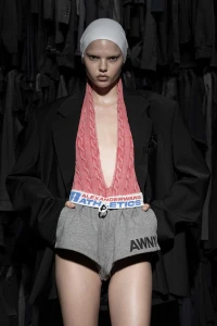 Model Winter wearing an oversized black blazer, pink cable-knit halter top, and grey athletic shorts against dark hanging garments.