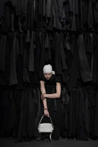 Model wearing a white headscarf and dark clothing standing against a dense backdrop of hanging black garments holding a white bag