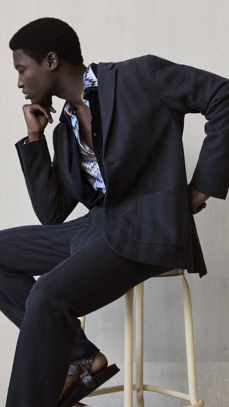 Black male fashion model in a dark blazer, patterned shirt, and pants is sitting on a stool in a studio with a neutral background.