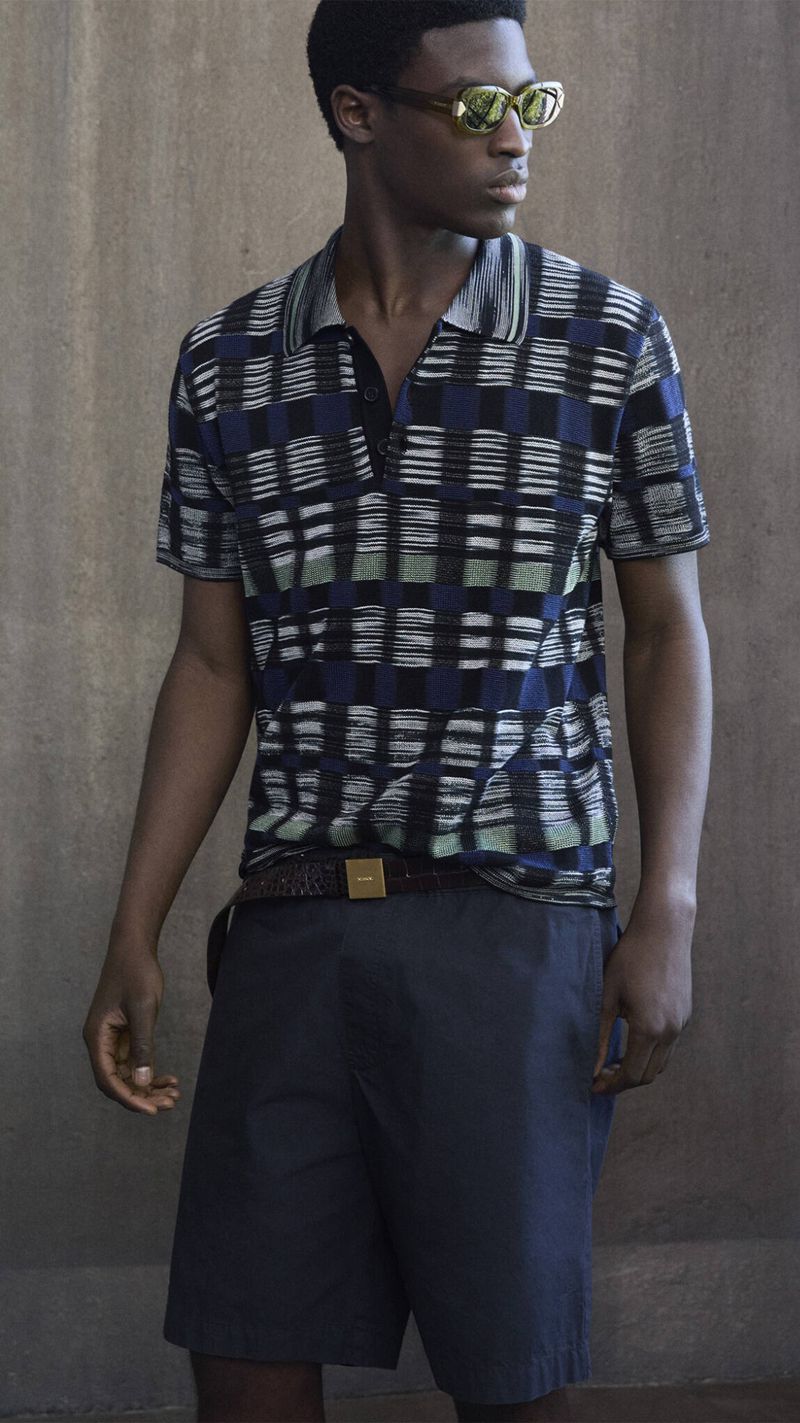 Young Black male model wearing sunglasses and a patterned short-sleeved shirt poses against a neutral backdrop in a fashion editorial shot.