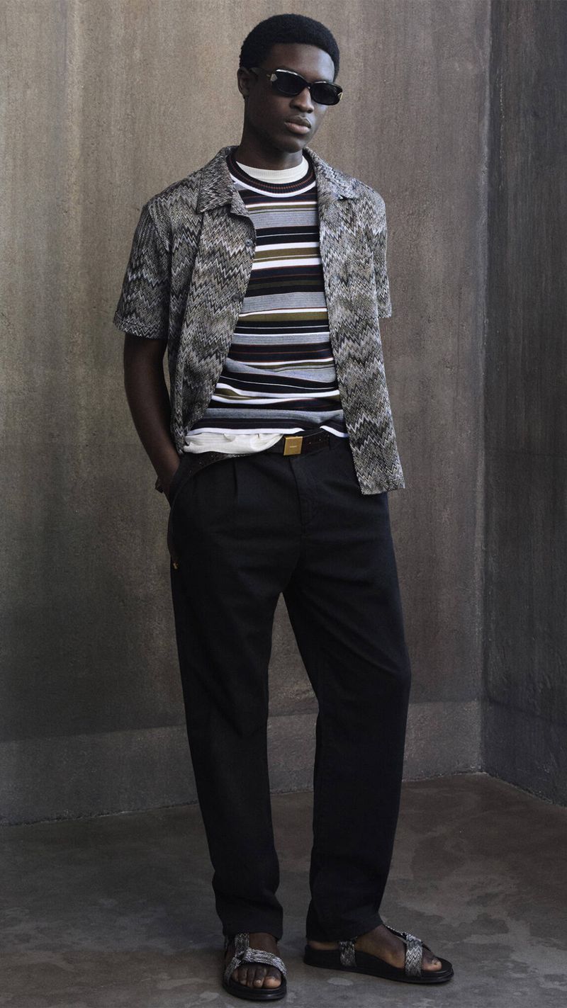 Fashionable adult Black male poses in open patterned shirt, striped tee, black pants, and sandals against a neutral textured background.