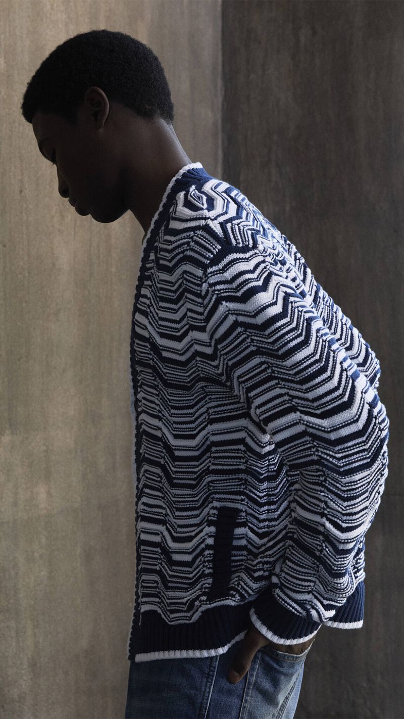 Man in patterned blue and white cardigan poses against a textured wall. He wears jeans in this medium shot fashion editorial image.