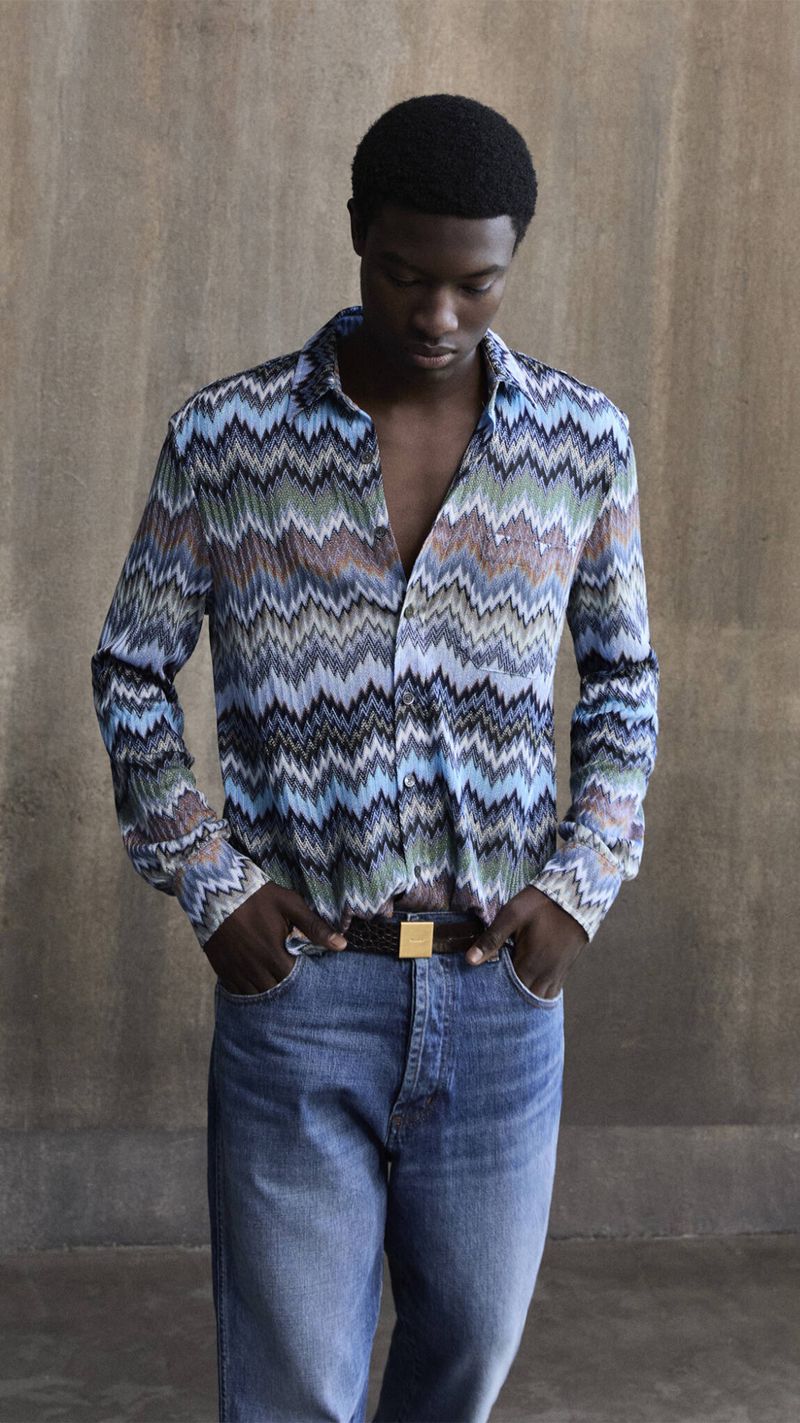 A dark-skinned male model poses indoors wearing a blue patterned button-down shirt and jeans, his hands casually placed in his pockets.