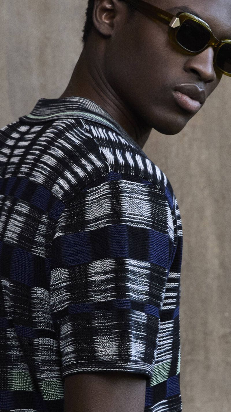 An adult male model with dark skin poses while wearing sunglasses and a polo shirt with a unique square pattern against a neutral background.