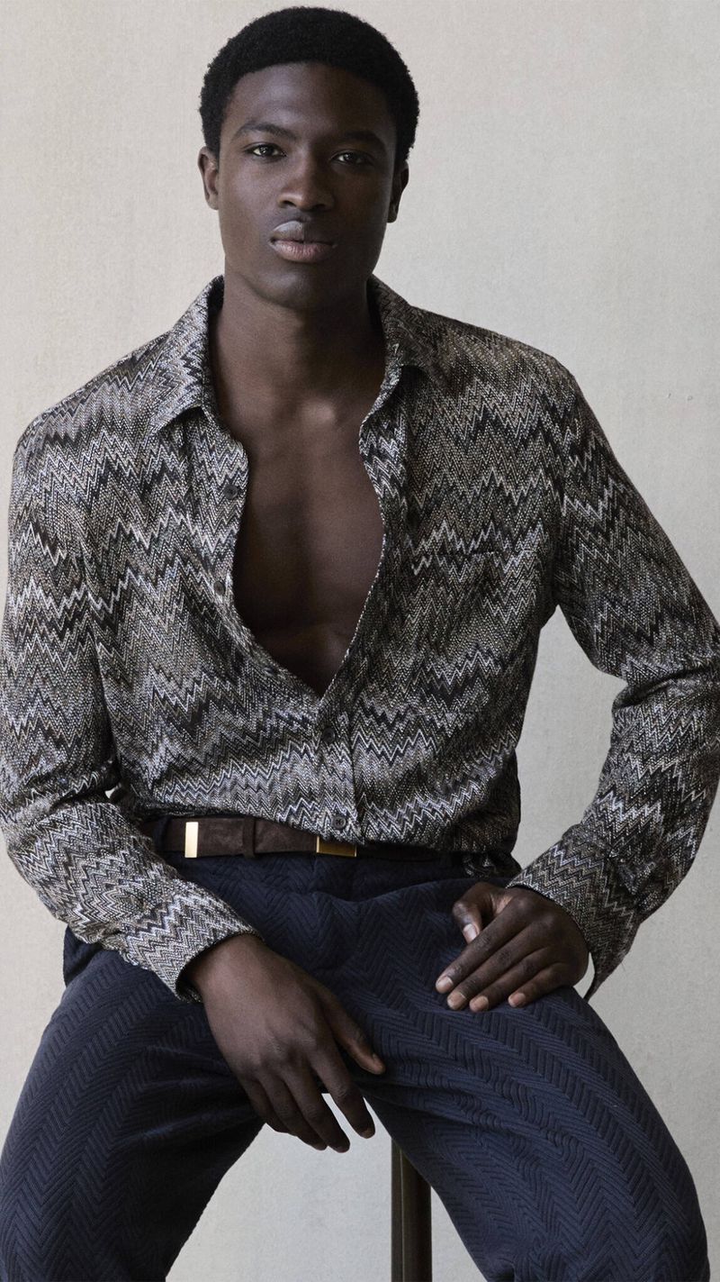 Portrait of a Black man seated on a stool in an indoor setting wearing a patterned shirt and dark pants looking at the camera.