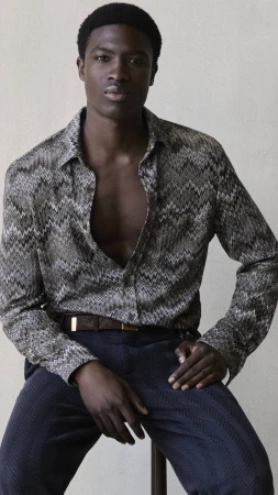 Model Momo Ciss wearing an open chevron patterned shirt and dark trousers seated against a neutral studio backdrop.