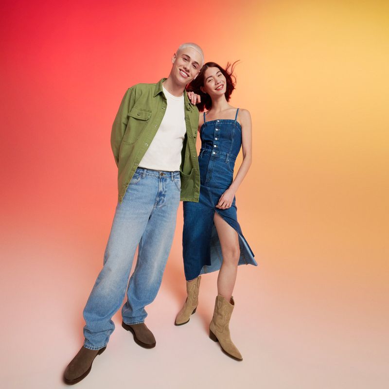 Stylish couple posing against a vivid gradient backdrop. The man wears a green shirt and jeans, while the woman wears a denim dress and boots.