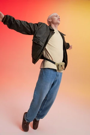 Young man with short bleached hair wearing a dark jacket, light t-shirt, and blue jeans leaping against a gradient background.