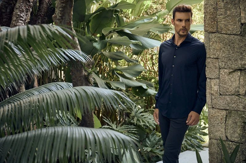 An adult male poses in a dark shirt and pants next to a stone wall, surrounded by tropical foliage and greenery in a natural outdoor setting.