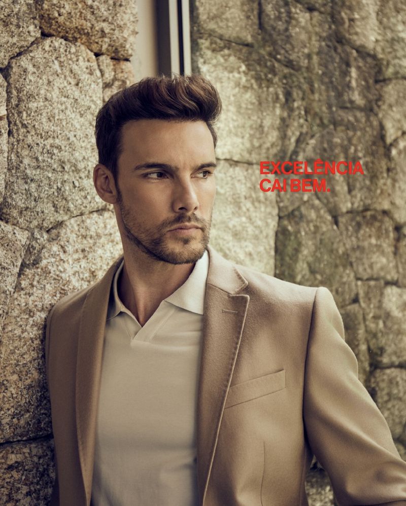 Portrait of an adult man with styled hair wearing a tan blazer and a white collared shirt, posing in front of a textured rock wall.