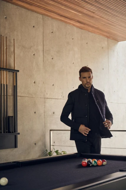 Man in a dark jacket standing next to a pool table inside a modern home interior with a concrete wall and wood ceiling detail.