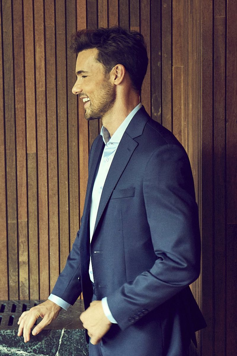 Profile view of a handsome adult male in a blue suit and light blue collared shirt smiling indoors with a wood-panelled background.