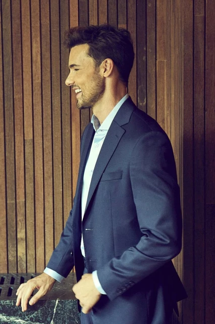 Profile view of a handsome adult male in a blue suit and light blue collared shirt smiling indoors with a wood-panelled background.