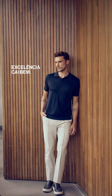 A man in a dark polo shirt and light pants leans against a wood wall, looking off to the side in a fashion style advertisement.