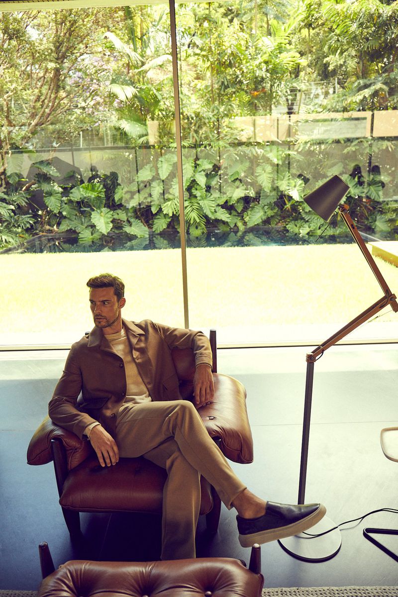 A casually dressed adult male sits in a leather chair in a light-filled indoor space with an expansive garden view.