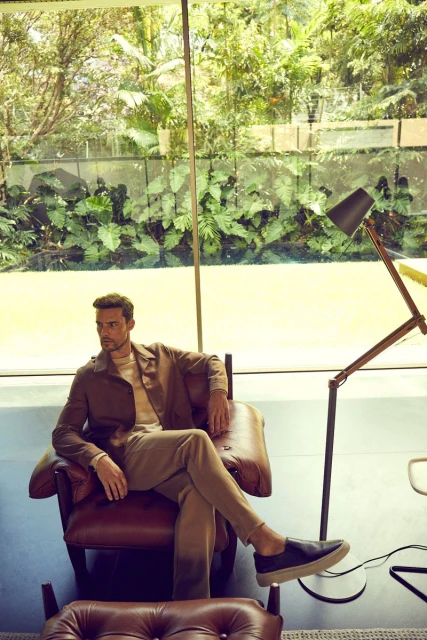 A casually dressed adult male sits in a leather chair in a light-filled indoor space with an expansive garden view.
