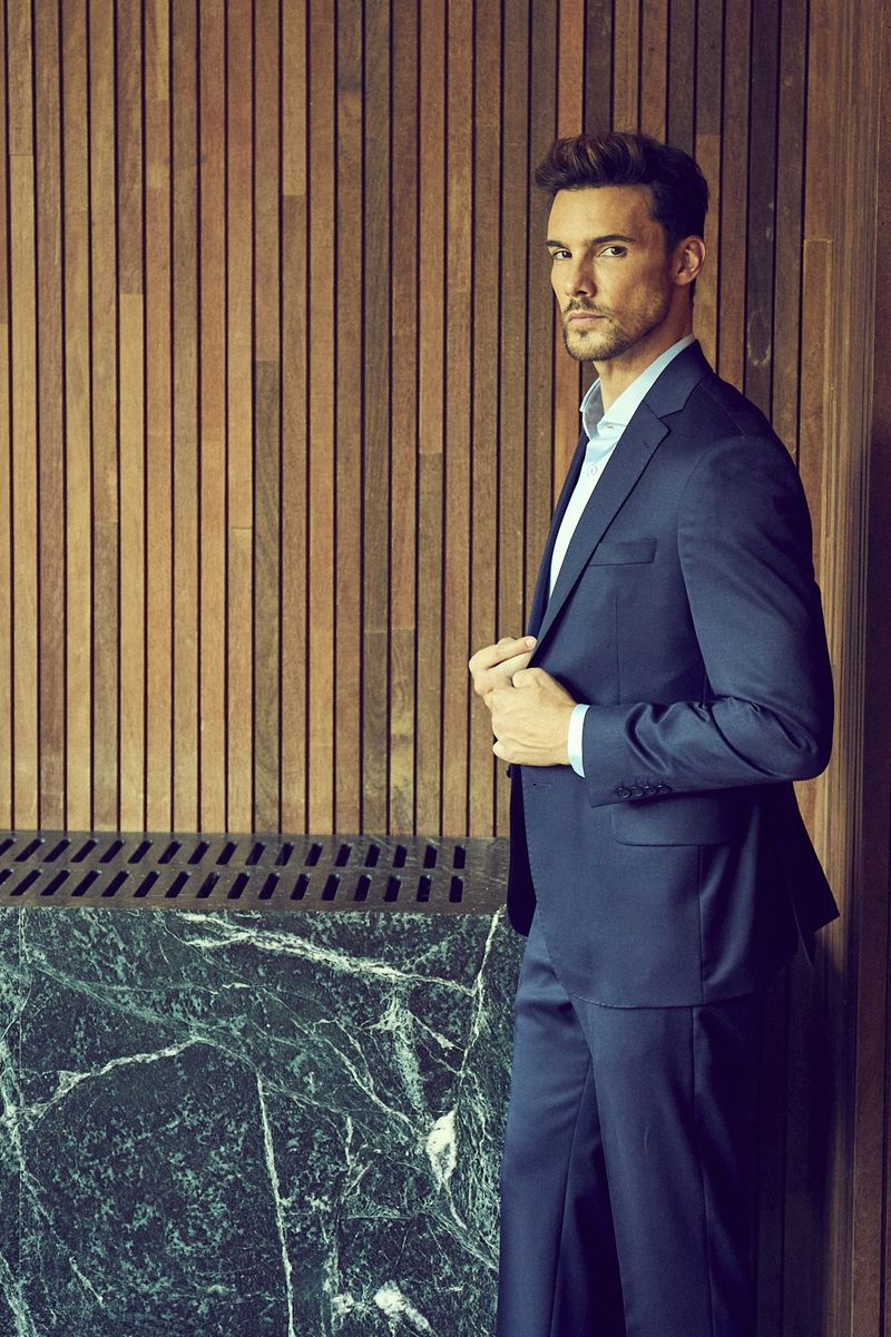 A stylish adult male in a blue suit poses against a wood panel wall with marble detail, bathed in soft, diffused lighting.