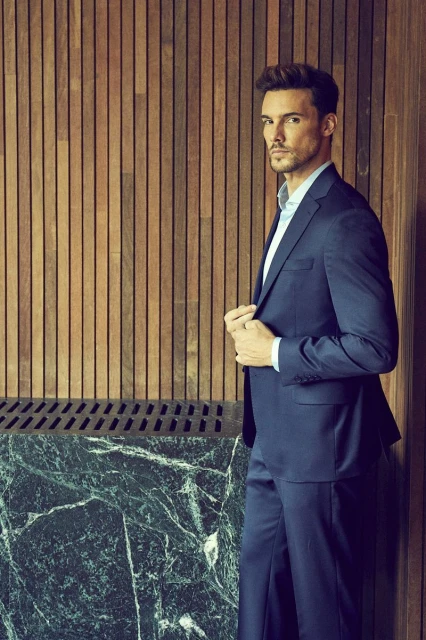 A stylish adult male in a blue suit poses against a wood panel wall with marble detail, bathed in soft, diffused lighting.