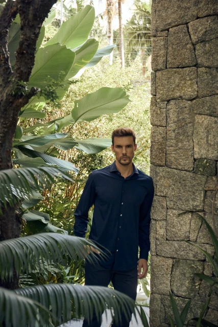Man in navy blue shirt and pants standing outdoors near stone wall with tropical greenery in background, posing in natural light.