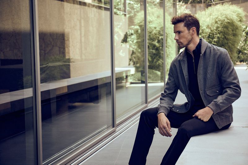 A stylish adult male in a gray jacket and black pants is sitting outside by large windows. He's looking to the side in a contemplative pose.
