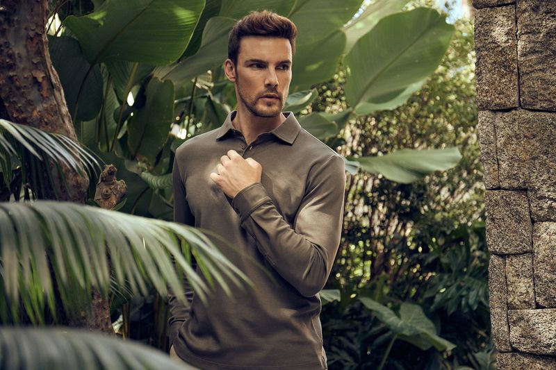 Portrait of a handsome adult male model wearing an olive green long-sleeved polo shirt in an outdoor tropical setting with lush vegetation.