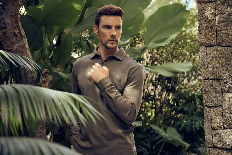 Portrait of a handsome adult male model wearing an olive green long-sleeved polo shirt in an outdoor tropical setting with lush vegetation.