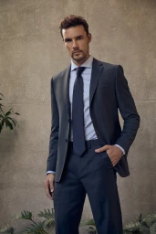Man posing in a dark charcoal tailored suit, light blue dress shirt, and navy tie against a neutral concrete wall with foliage.